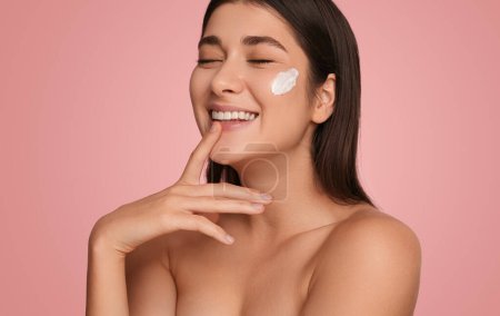 Photo for Beautiful young female with closed eyes smiling and touching chin while applying cream on face on pink background in studio - Royalty Free Image