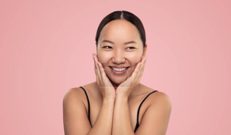Téléchargez les photos : Positive young ethnic female smiling and touching face while looking at copy space isolated on pink background in studio - en image libre de droit