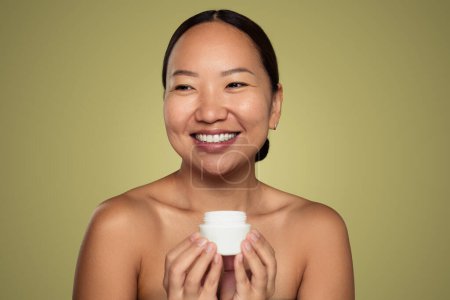Photo for Cheerful young ethnic female with clean skin smiling and holding a jar of skincare cream while standing against green background - Royalty Free Image