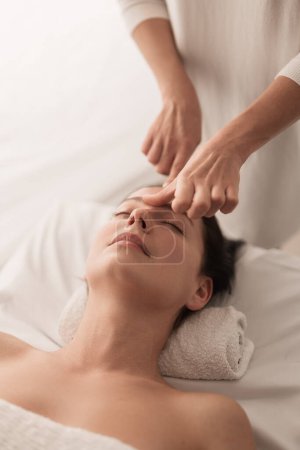 Foto de High angle of crop anonymous masseur massaging forehead of young female client lying on table with rolled towel, during beauty procedure in light spa salon - Imagen libre de derechos