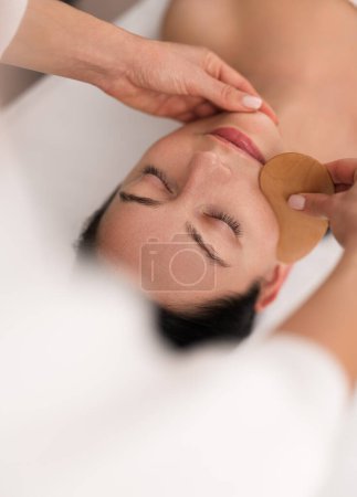 Foto de From above of crop unrecognizable beautician massaging face of calm young woman, lying on couch with closed eyes with wooden gua sha in spa salon - Imagen libre de derechos