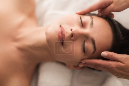 Téléchargez les photos : Top view of relaxed young female client with dark hair and perfect skin lying on massage table with closed eyes, during beauty procedure in modern light salon - en image libre de droit