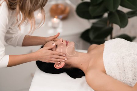 Téléchargez les photos : From above side view of crop unrecognizable female cosmetician touching face of client lying on massage table during beauty procedure in modern spa salon - en image libre de droit