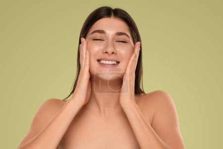 Téléchargez les photos : Joyful young female millennial with bare shoulders and long dark hair, smiling happily with closed eyes and touching cheeks while applying moisturizing cream on face against green background - en image libre de droit