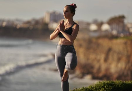 Téléchargez les photos : Young barefoot female in leggings and bra clasping hands and meditating in Tree pose, during yoga session on blurred background of waving sea and cliff - en image libre de droit