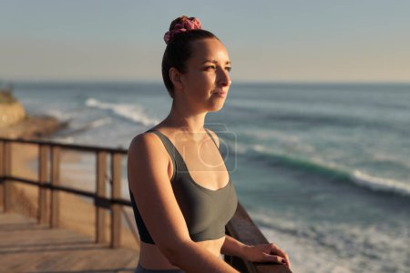 Téléchargez les photos : Female athlete in black bra looking away and observing waving sea while taking break in fitness training on terrace at sunset - en image libre de droit