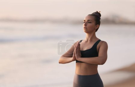 Téléchargez les photos : Relaxed young fit female with dark hair in activewear standing on sandy beach near ocean with namaste hands, while meditating with closed eyes at sunset - en image libre de droit