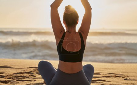 Téléchargez les photos : Back view of unrecognizable slim female in sportswear sitting on sandy beach near waving sea during fitness training - en image libre de droit
