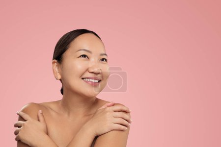 Téléchargez les photos : Optimistic Korean female with perfect skin looking away with smile and touching bare shoulders during spa session against pink background - en image libre de droit