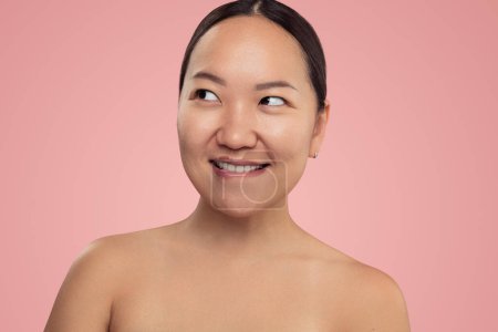 Téléchargez les photos : Curious young Asian female model with perfect skin and bare shoulders biting lip and looking away against pink background - en image libre de droit