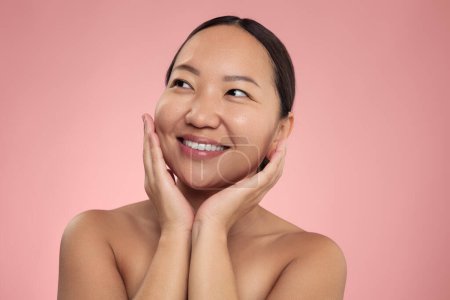 Téléchargez les photos : Delighted young Asian female model with dark hair and perfect skin, smiling and looking away while touching cheeks after skin care procedure against pink background - en image libre de droit