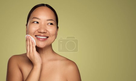 Téléchargez les photos : Cheerful young asian female touching clean skin with reusable makeup disk, while looking to the side against green background with copy space - en image libre de droit