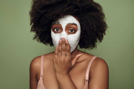 Foto de Surprised young African American female millennial with long dark curly hair in lingerie, covering mouth with hand and looking at camera while standing against green background with mask on face - Imagen libre de derechos