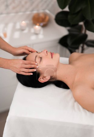 Téléchargez les photos : From above of crop unrecognizable masseuse doing gentle face massage to relaxed female client with long dark hair, lying on table with closed eyes during spa procedure in modern salon - en image libre de droit