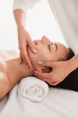 Foto de Crop unrecognizable cosmetician doing anti aging face massage to young content female client with dark hair, lying on couch with rolled tower in modern light beauty salon - Imagen libre de derechos
