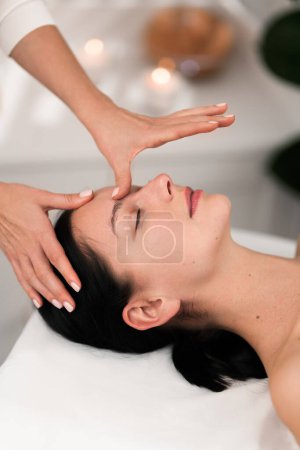 Téléchargez les photos : High angle of crop unrecognizable therapist pressing on forehead of calm young female customer, lying on massage table with closed eyes during acupressure treatment in modern salon - en image libre de droit