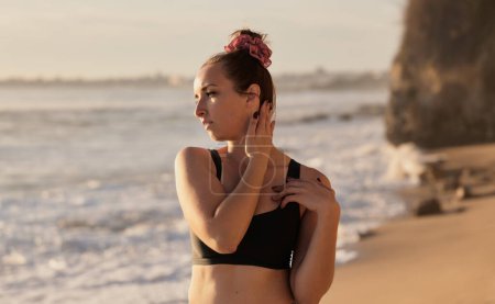 Foto de Vista lateral de la joven atleta en ropa deportiva de pie en la playa de arena cerca del mar ondulante y mirando hacia otro lado - Imagen libre de derechos