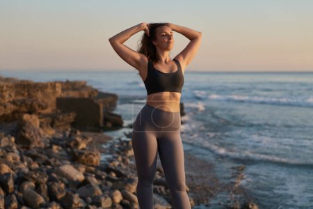Foto de Self assured young fit woman in activewear touching long wavy hair, while standing in rocky beach near ocean with closed eyes after practicing yoga at sunset - Imagen libre de derechos