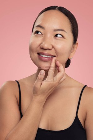 Téléchargez les photos : Glad asiatique femelle regarder loin avec sourire et barbouiller baume hydratant sur les lèvres et regarder loin sur fond rose - en image libre de droit