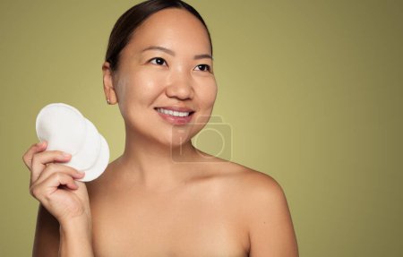 Photo for Happy Asian female with bare shoulders demonstrating bunch of clean reusable pads and looking away with smile during skin care routine against olive background - Royalty Free Image