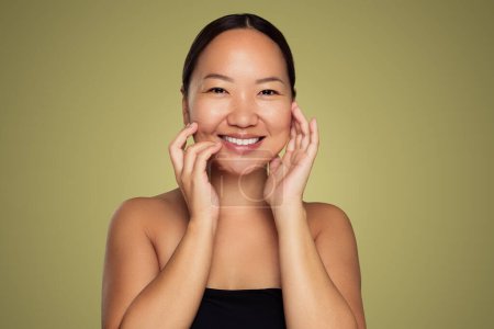 Téléchargez les photos : Jeune ethnique asiatique femelle avec naturel propre regard touchant visage et souriant tout en se tenant debout sur fond vert - en image libre de droit