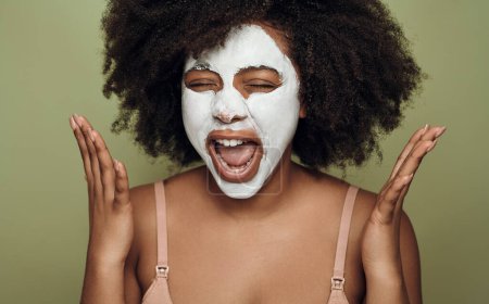 Foto de Expressive young African American female with dark curly hair and white face mask screaming with closed eyes during skin care routine against green background - Imagen libre de derechos