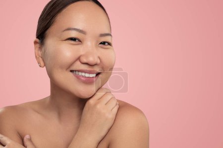 Téléchargez les photos : Cheerful young ethnic female millennial with perfect skin and bare shoulders wearing no makeup smiling and looking at camera against pink background - en image libre de droit