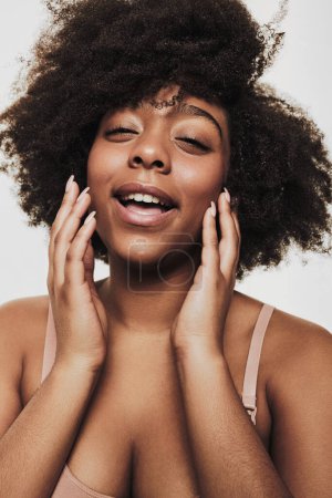 Téléchargez les photos : Young laughing African American woman touching soft skin on cheeks and looking at camera on white background - en image libre de droit