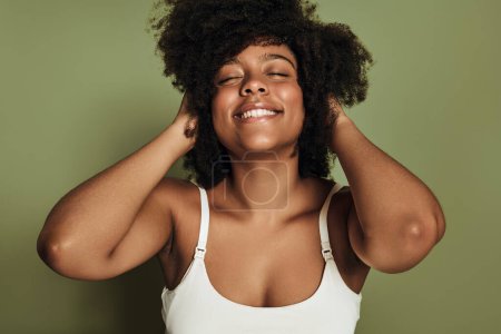 Téléchargez les photos : Joyeux jeune afro-américaine millénium féminin en soutien-gorge blanc souriant joyeusement avec les yeux fermés et toucher les cheveux noirs bouclés sur fond vert - en image libre de droit