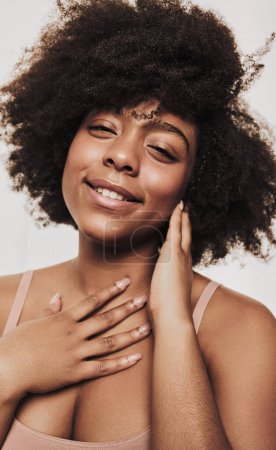 Téléchargez les photos : Positive young African American woman with curly hair touching chest and neck and looking at camera with smile during beauty routine against gray background - en image libre de droit