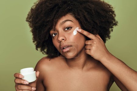Photo for Concentrated young African American female millennial with dark curly hair and bare shoulders, applying moisturizing cream on face and looking at camera against green background - Royalty Free Image