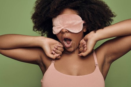 Photo for Sleepy young African American female with dark curly hair in too and eye mask yawning after awakening against green background - Royalty Free Image