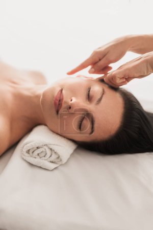 Foto de From above of relaxed young female client with long dark hair and bare shoulders lying on massage table with closed eyes, during skincare treatment in light salon - Imagen libre de derechos