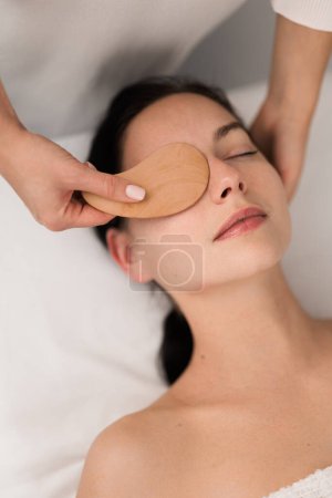 Photo for From above of crop unrecognizable cosmetician using wooden gua sha while massaging face of female client during beauty procedure in modern salon - Royalty Free Image