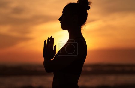 Téléchargez les photos : Vue latérale silhouette de jeune femelle tenant la main dans la pose namaste et priant au coucher du soleil à l'océan - en image libre de droit