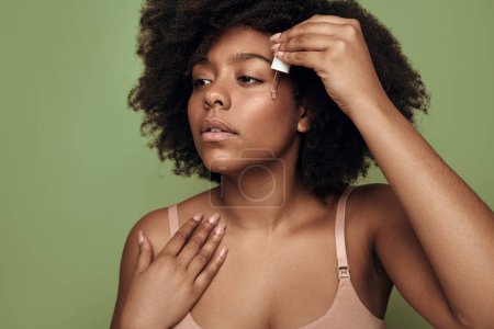 Téléchargez les photos : Black woman in beige bra with curly hair touching shoulder and looking away, while applying moisturizing serum on cheek during skin care routine against green background - en image libre de droit