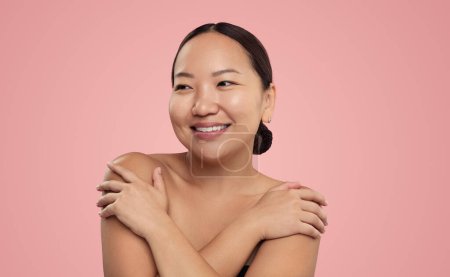 Photo for Young ethnic female with clean glowing skin embracing bare shoulders and looking away with smile on pink background - Royalty Free Image