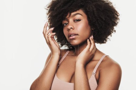Téléchargez les photos : Femme afro-américaine avec la coiffure afro toucher le visage et en regardant la caméra sur fond blanc montrant visage propre et concept de soins de la peau - en image libre de droit
