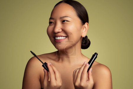 Foto de Gleeful young Korean woman with bare shoulders showing mascara and looking away with smile against olive background - Imagen libre de derechos