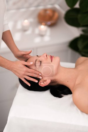 Photo for High angle of crop beautician rubbing temples of relaxed client with closed eyes during face massage session in spa salon - Royalty Free Image
