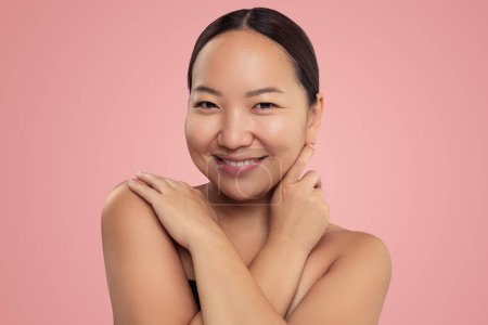 Téléchargez les photos : Positive young ethnic female with bare shoulders smiling and touching face against pink background - en image libre de droit