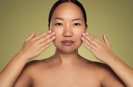 Téléchargez les photos : Confident young Asian female model with perfect skin bare shoulders and dark hair, touching face while applying moisturizing cream and looking at camera against green background - en image libre de droit