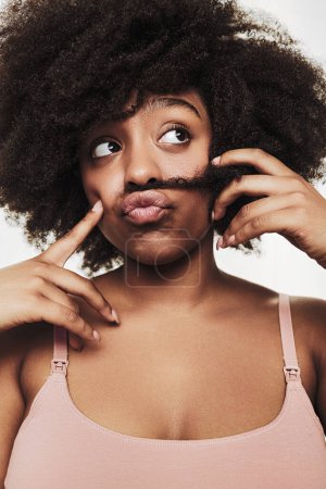 Foto de Thoughtful young African American female model in bra pouting lips and making mustache with dark curly hair while looking away against white background - Imagen libre de derechos