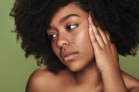 Téléchargez les photos : Jeune femme afro-américaine aux cheveux bouclés touchant le visage et regardant loin sur fond vert. Concept beauté et peau propre - en image libre de droit
