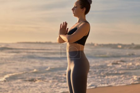 Téléchargez les photos : Vue latérale de la culture paisible jeune femme en forme avec les cheveux foncés dans les vêtements de sport méditant avec les yeux fermés, tout en effectuant Montagne avec les mains de prière posent pendant la séance de yoga près de l'océan ondulé - en image libre de droit