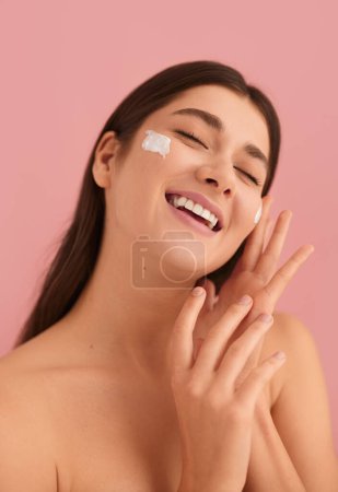 Foto de Delighted young female millennial with dark hair and bare shoulders, smiling happily with closed eyes while applying moisturizing cream on face during during skin care routine against pink background - Imagen libre de derechos