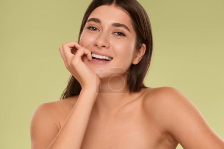 Photo for Closeup of beautiful young female with perfect skin and bare shoulders smiling and looking at camera while standing on green background in studio - Royalty Free Image