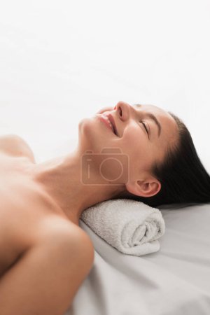 Téléchargez les photos : High angle of delighted young female with bare shoulders and long dark hair, lying on massage table with closed eyes and smiling during spa session in light salon - en image libre de droit