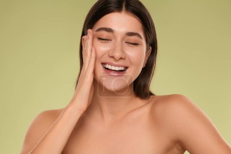 Photo for Cheerful young brunette with bare shoulders smiling with closed eyes and touching soft cheek during skin care routine against green background - Royalty Free Image
