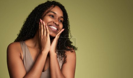 Téléchargez les photos : Femme afro-américaine optimiste avec des cheveux bouclés touchant le visage et regardant l'espace de copie tout en se tenant debout sur fond vert en studio - en image libre de droit